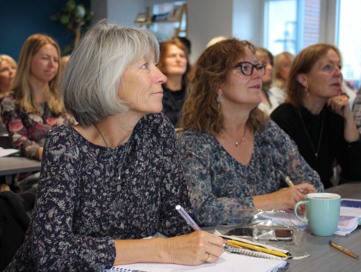 Two women sitting at a table with notebooks and pens, listening attentively in a room with other people.